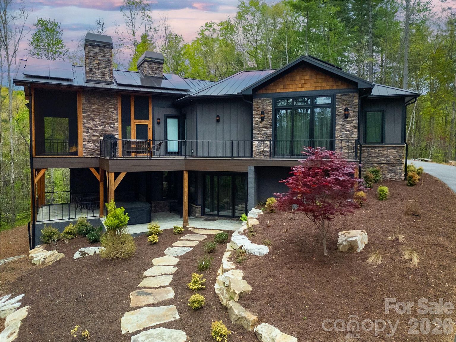 The rear elevation at twilight. Two stone chimneys rise from a black metal roof. A covered upper balcony, a glass-and-steel railing, a Japanese maple in front of dry-stacked stone, and a stone path picking its way down through the landscape.