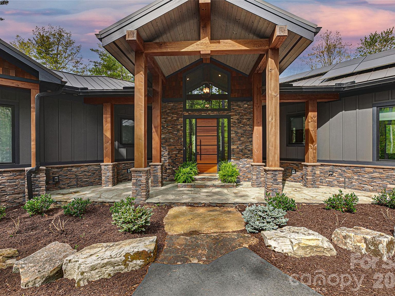 The entry portico. A timber gable on heavy posts, dry-stacked stone columns, a wood front door behind a smaller stone landing. Stone path bedded into the rock garden, twilight sky overhead.