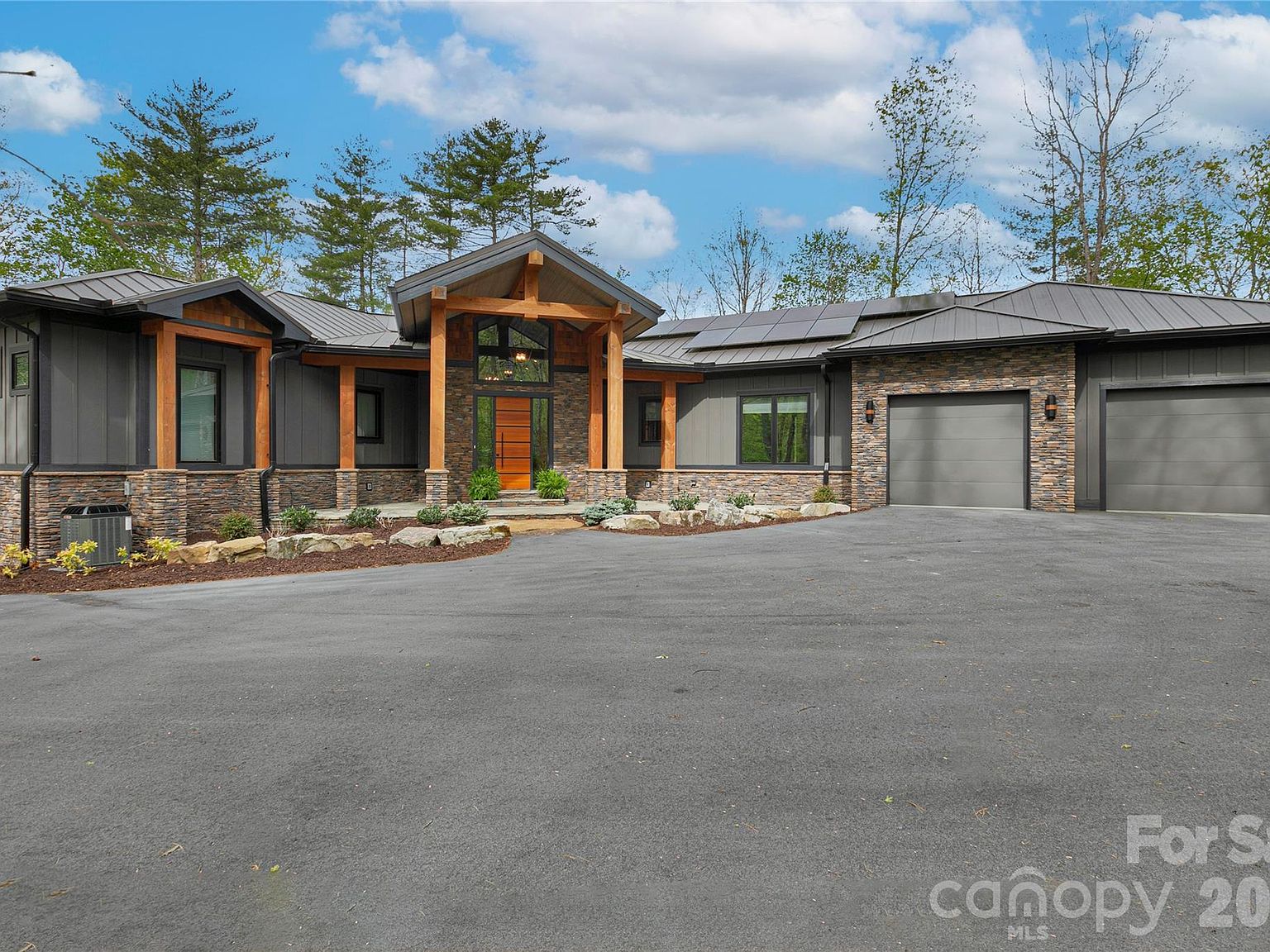 The front elevation by morning. Board-and-batten siding in deep charcoal, timber columns over the portico, two garage doors, a calm gravel drive. Spring sky in clean blue overhead.