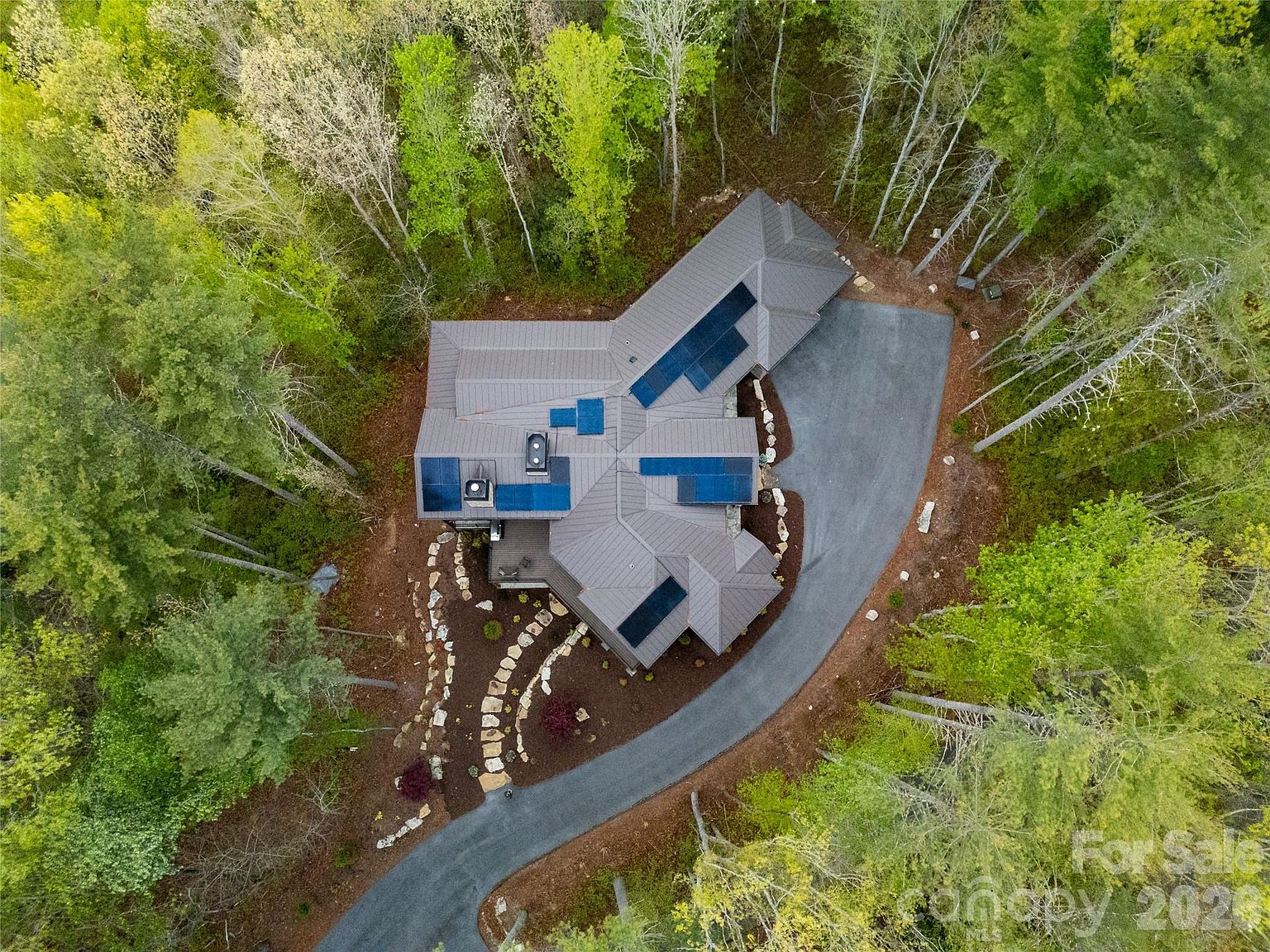 An aerial of the property. The home set on a clearing in dense hardwood forest, the driveway curving away through the trees, slate-blue roofs, a stone retaining wall along the rear.